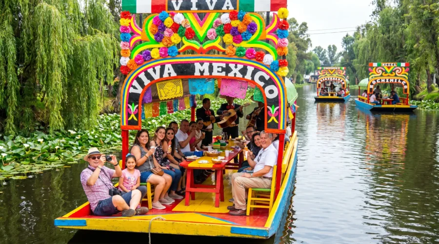 xochimilco-food-tour-by-mexico-foodie-tours Colorful trajinera decorated with flowers and papel picado gliding through the canals on the Xochimilco Tour by Mexico Foodie Tours, with mariachi musicians and guests aboard.